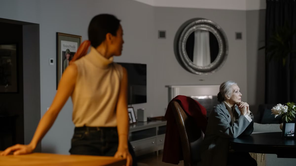 Senior woman deep in thought at a table with a younger woman in focus background, evoking a quiet, introspective mood.