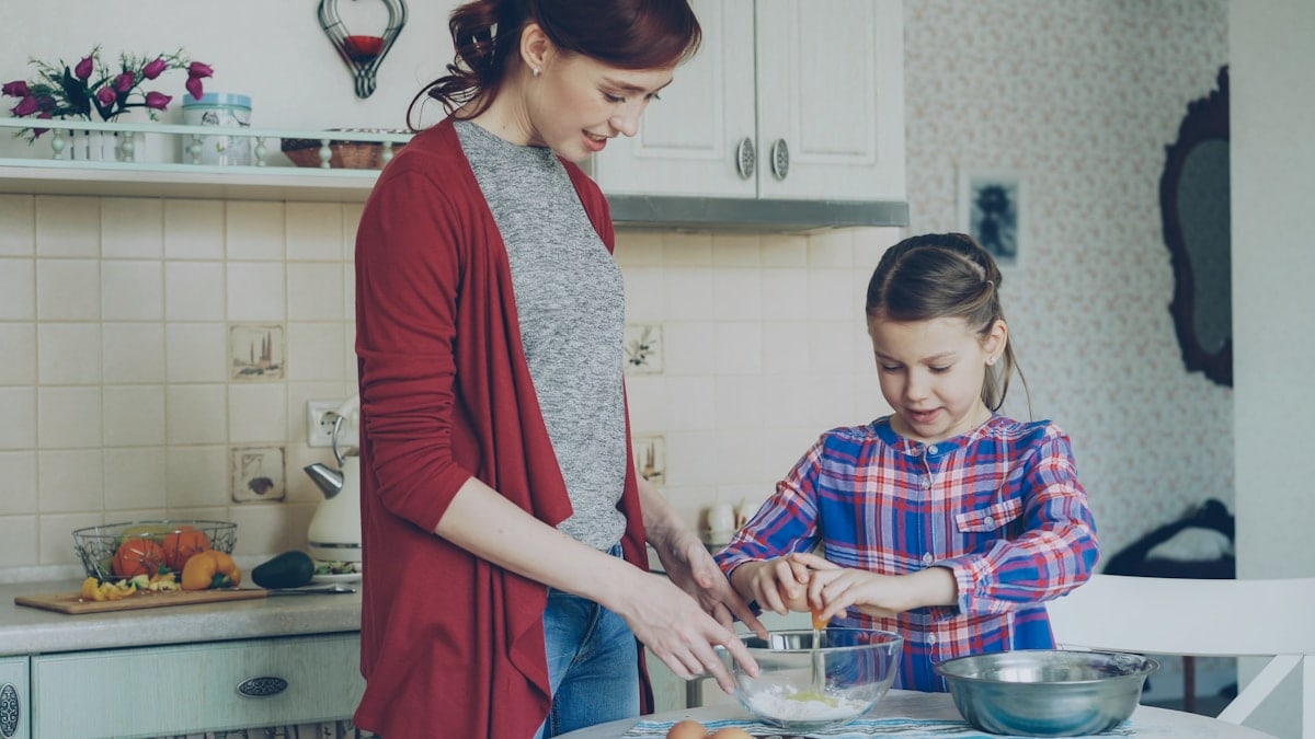 Little cheerful girl helping her mother in the kitchen cropping egg into bowl and mixing dough for cookies. Family, food, home and people concept